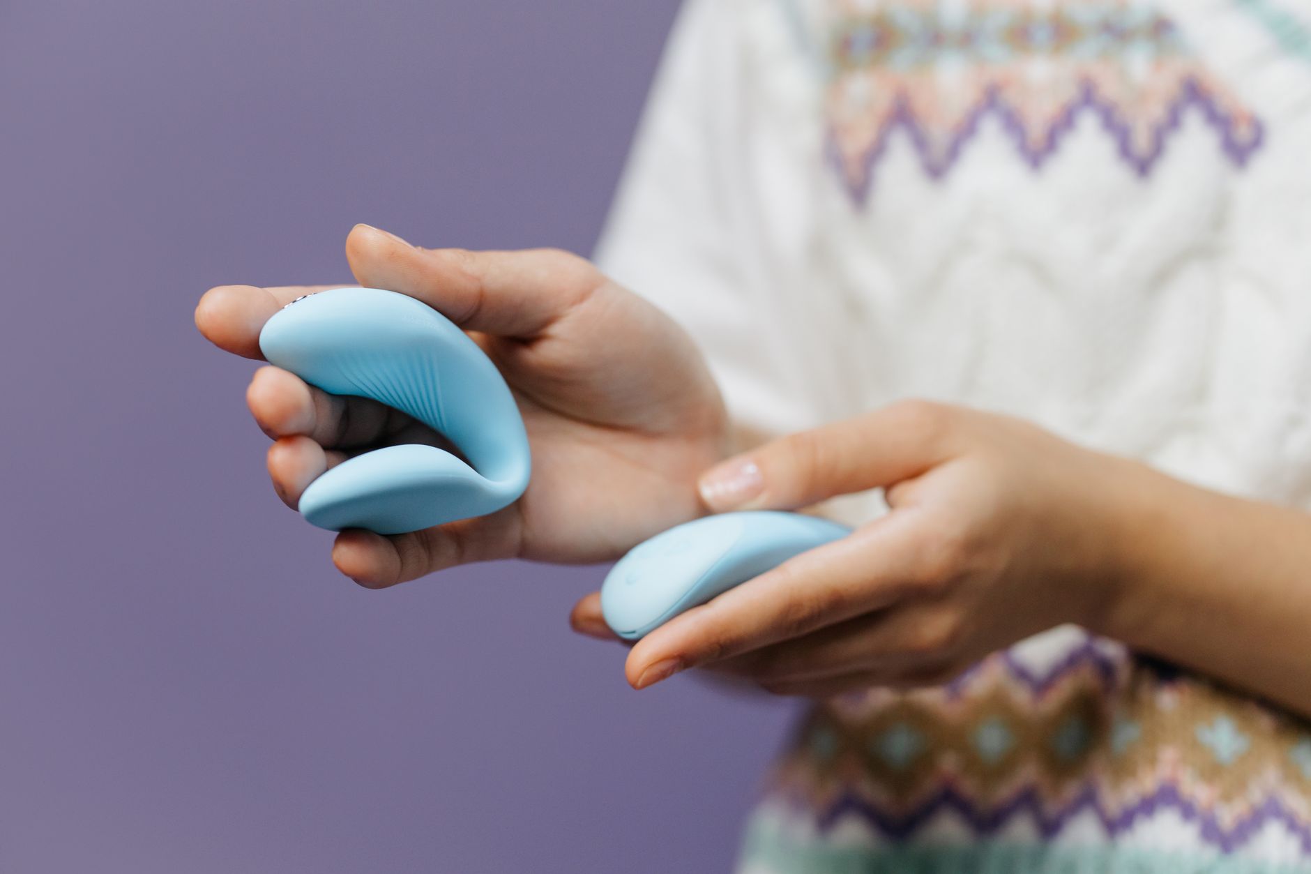 Hands holding a sleek blue clitoral vibrator against a purple background